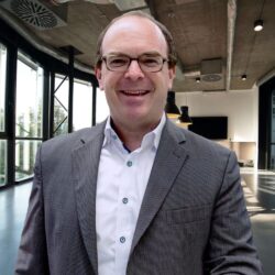 A man with glasses, wearing a gray suit jacket and white shirt, is smiling. He is standing in a modern, industrial-style office with large windows and exposed ductwork. Black pendant lights can be seen in the background, reflecting the sleek design typical of commercial juicer brands like Citrus America.