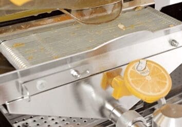 Close-up of a commercial citrus juicer with a fresh orange half positioned for juicing. The machine features a metal straining surface with juice remnants and a yellow lever handle. The juicer dispenses orange juice through a spout at the bottom.