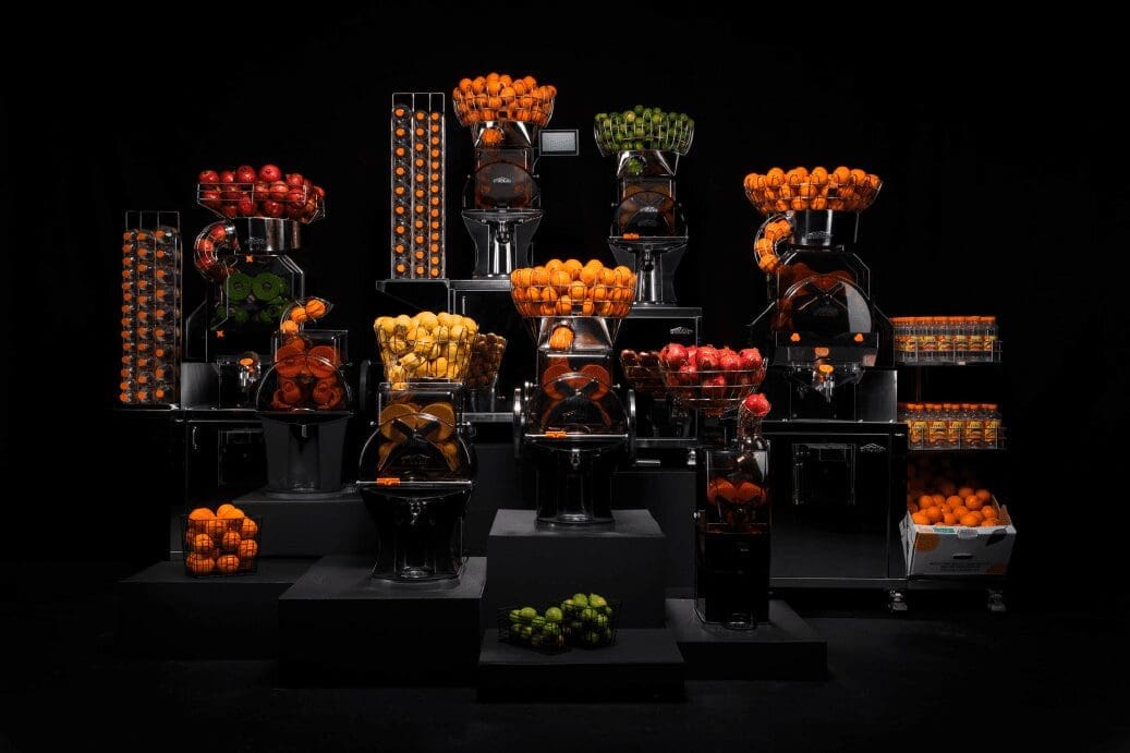 A display of various commercial juicers arranged in tiers against a black background. Each machine is filled with citrus fruits like oranges and apples, ready for juicing, with some stacked in clear containers and others at the base, creating a vibrant and colorful arrangement.