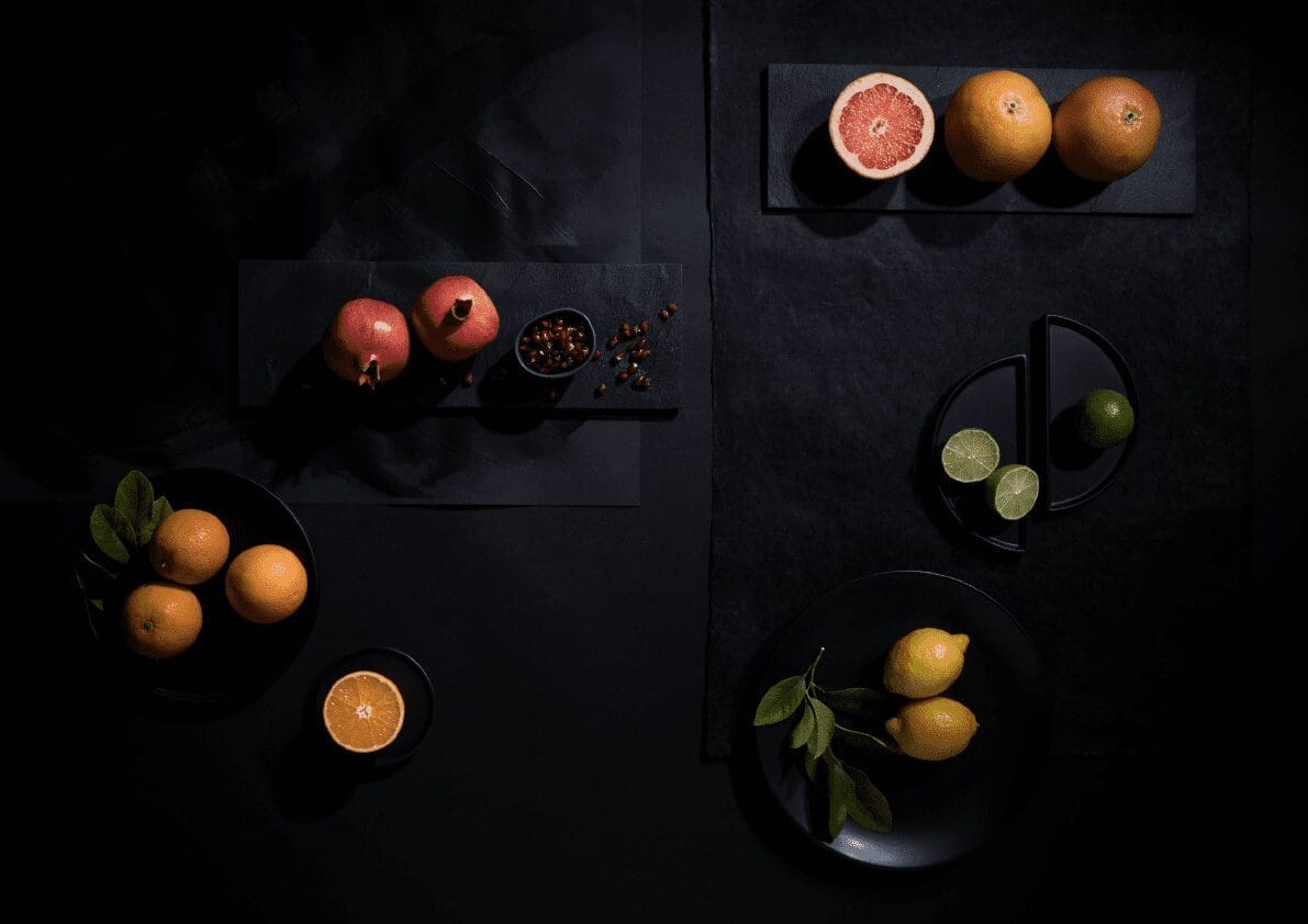 A dark, moody photo showcasing various citrus fruits. There are two pomegranates, halved grapefruit, oranges, limes, and lemons with leaves, all artistically arranged on black plates and slate boards against a dark background, creating a striking contrast reminiscent of a commercial juicer advertisement.