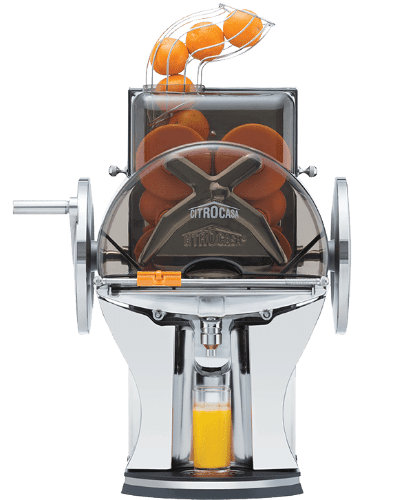 A chrome and black electric citrus juicer from Citrus America features a transparent compartment holding whole oranges. Oranges are being juiced, and fresh juice flows into a clear glass positioned underneath. A hand crank is visible on the left side of this commercial juicer.