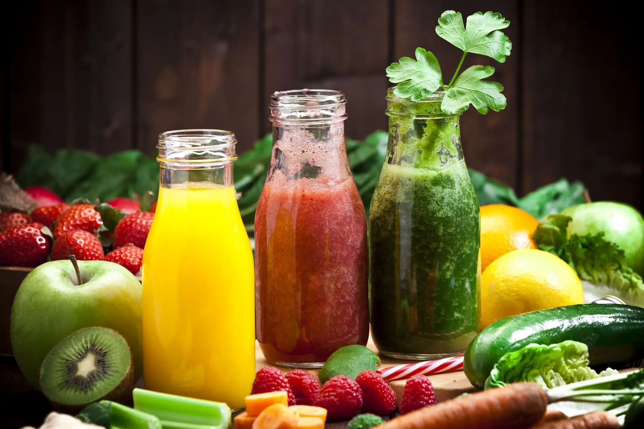 Three glass bottles filled with yellow, red, and green juices—our signature products—are surrounded by fresh fruits and vegetables like apples, strawberries, lemons, celery, carrots, and leafy greens.