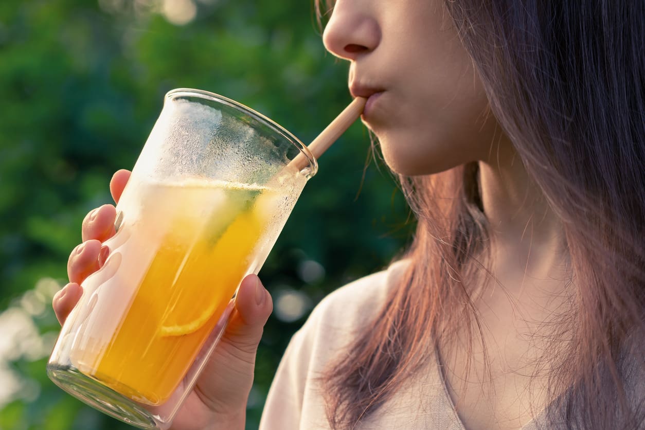 A person with long hair sips a refreshing-looking citrus drink with ice cubes and a slice of lime through a straw from a clear glass, set against a blurred green outdoor background.