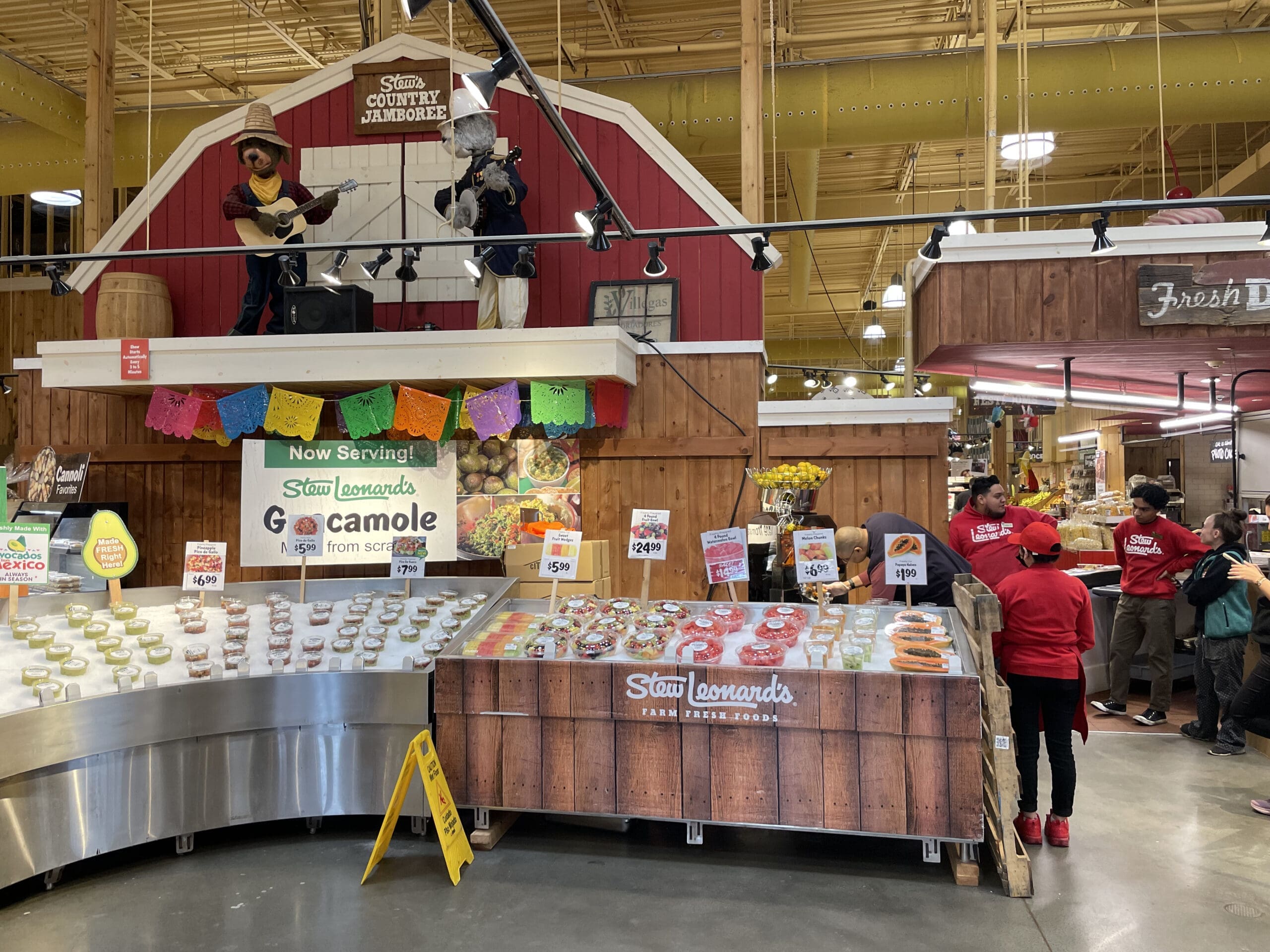 A store display promotes Staun Leonard's guacamole with various guacamole containers on ice next to a commercial juicer highlighting Citrus America products. Decor includes colorful banners, farm-themed cutouts, and signs with the prices and promotions. Several staff members in red shirts assist customers in the background.