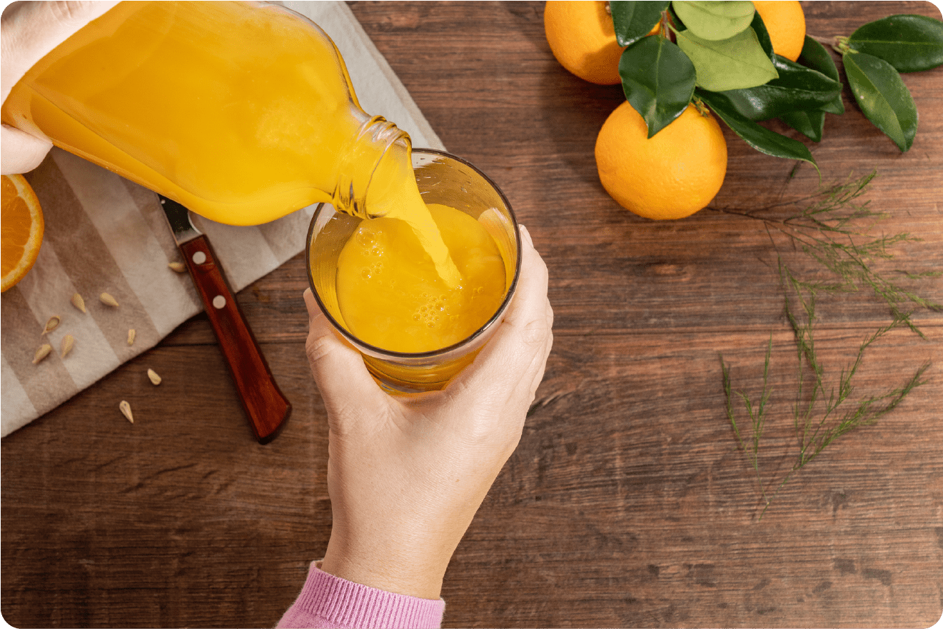 A person pours fresh orange juice from a glass bottle into a clear glass on a wooden table. Nearby, there are whole oranges with green leaves, an orange half, seeds, and a knife on a cloth, showcasing the vibrant bounty of Citrus America.