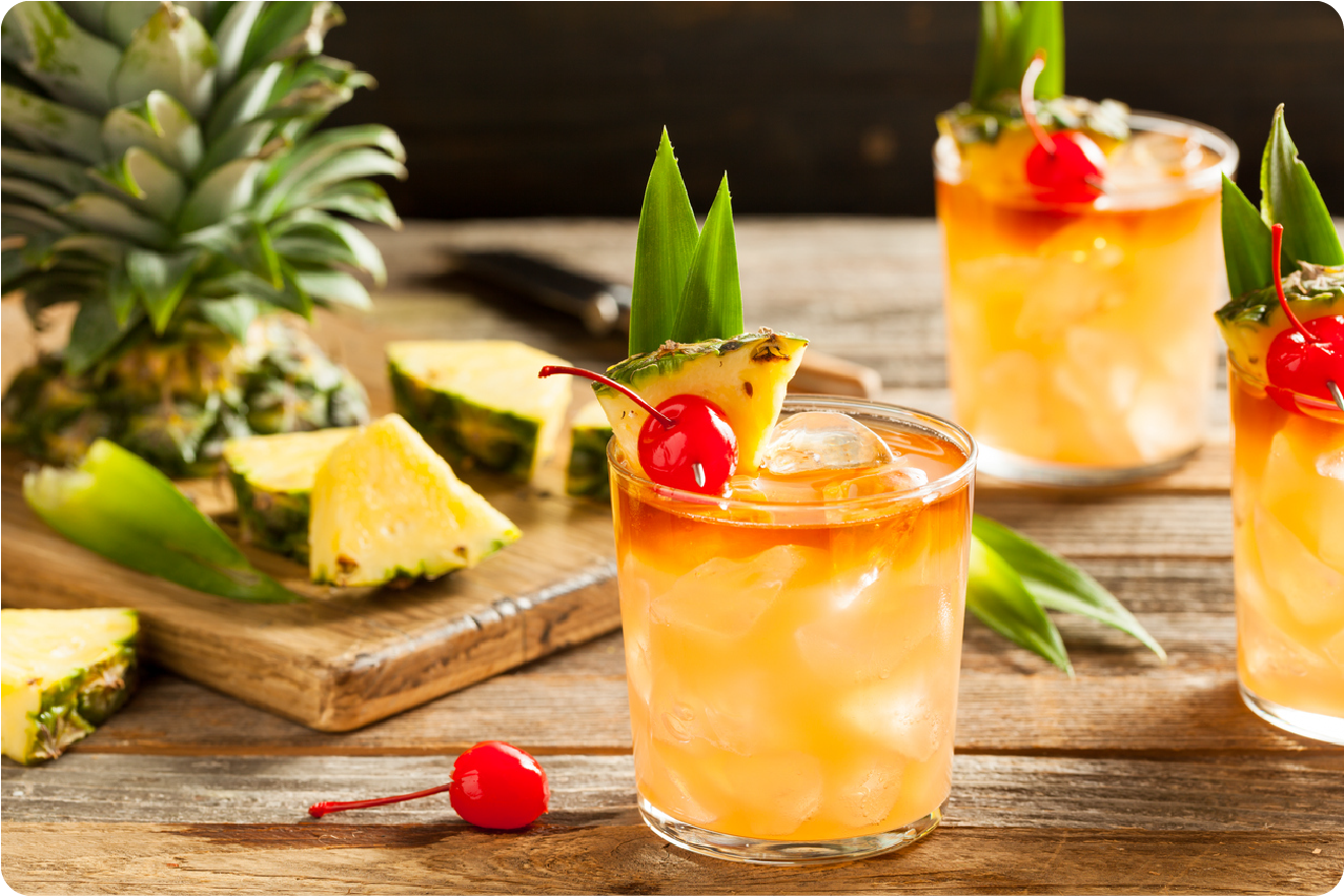 A refreshing tropical cocktail garnished with a pineapple slice, leaf, and cherry is served on a wooden table. In the background, more cocktails and a chopped pineapple with slices can be seen on a cutting board next to a Citrus America juicer.