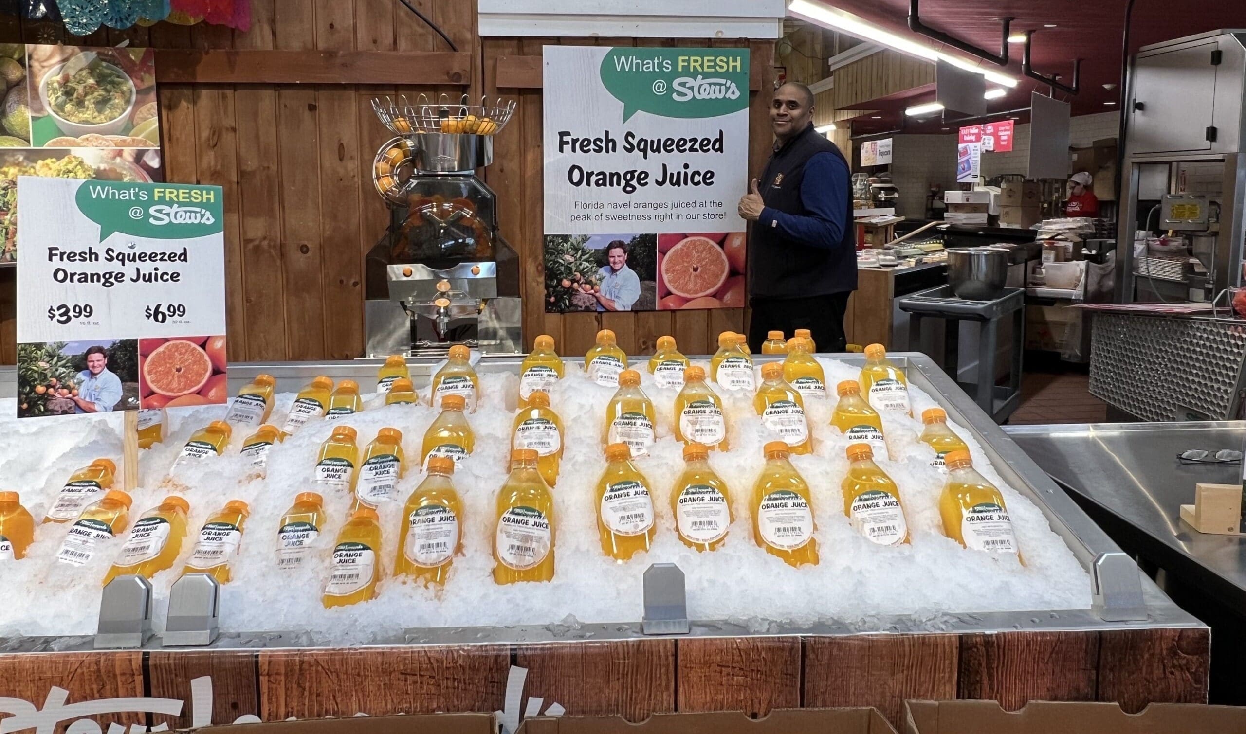 A man stands smiling near a display of fresh-squeezed orange juice bottles sitting on ice at a market. Signs above the citrus-themed display advertise the fresh orange juice for $3.99. The market area is surrounded by wooden decor and food preparation equipment, including a commercial juicer in the corner.