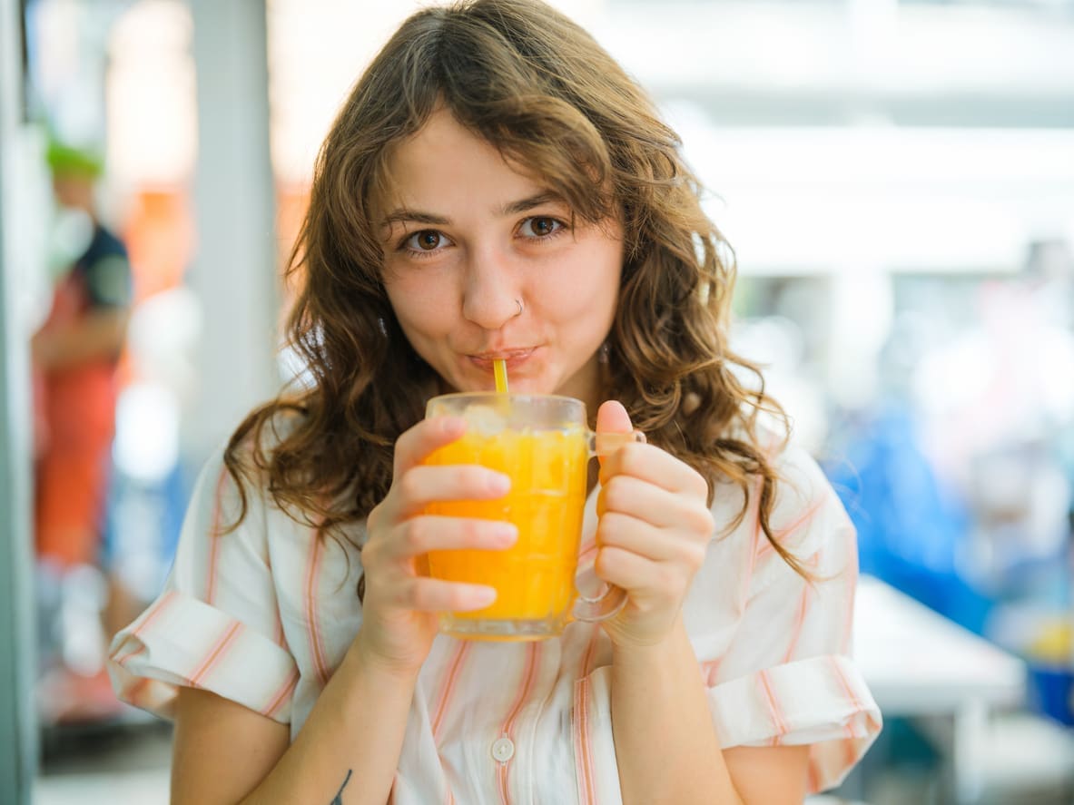 A young woman with wavy brown hair is sipping orange juice made from a commercial juicer through a straw. She is wearing a light-colored short-sleeved shirt, looking directly at the camera with a gentle smile. The background, featuring bright colors and shapes, is softly blurred.