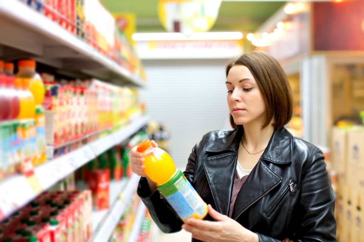 A woman with shoulder-length hair wearing a black leather jacket is standing in a grocery store aisle. She is examining a bottle of citrus orange juice among several colorful juice bottles on the shelves. The background shows more products and fluorescent lighting.