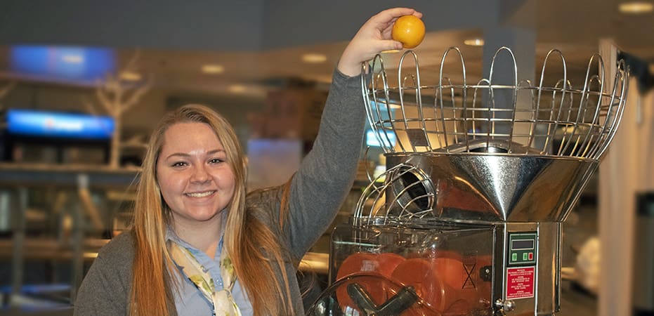 A woman with long blonde hair is smiling and placing an orange into a commercial citrus juicer. The machine, equipped with a wire basket for holding oranges, is positioned on a countertop in a well-lit room. The background includes blurred details of a kitchen or café setting.