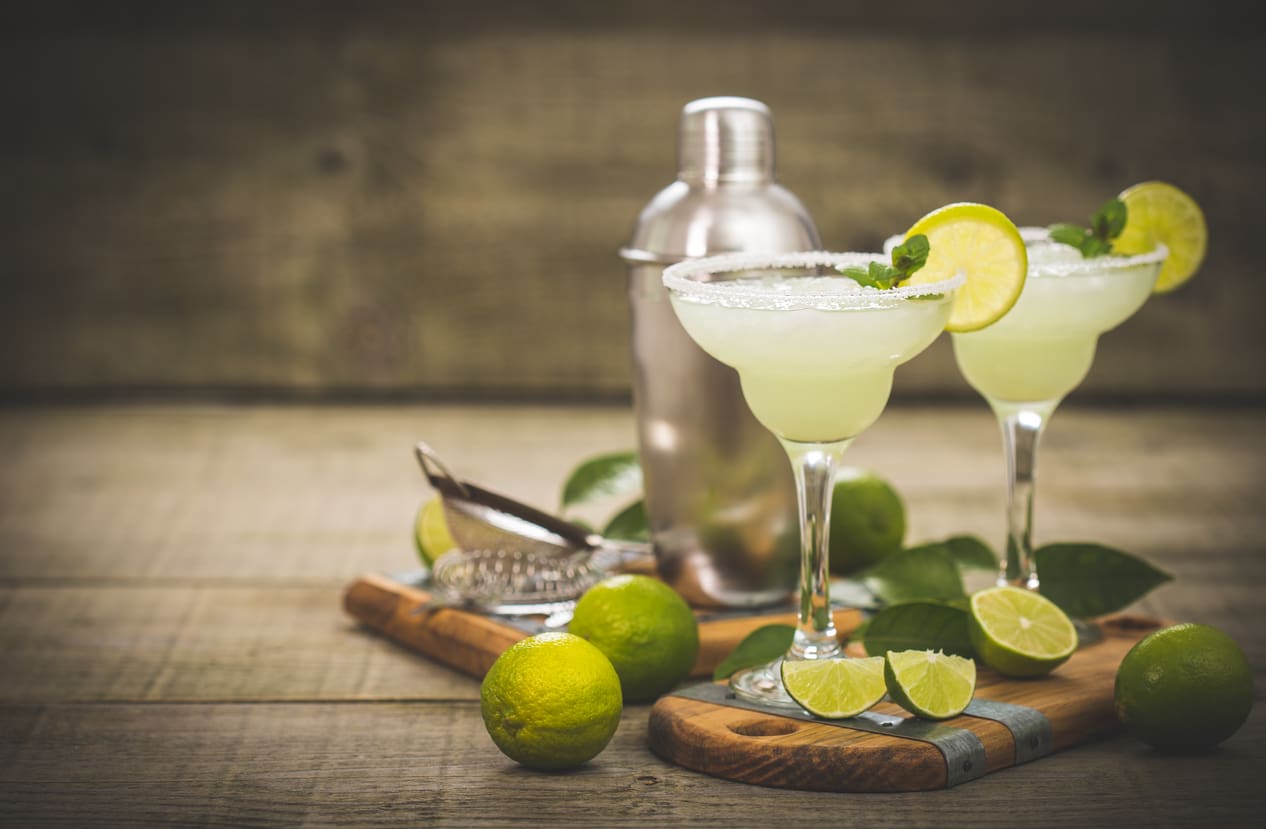 Two margarita glasses filled with a lime cocktail, each garnished with a lime slice and mint leaves, are placed on a wooden board. Fresh lime halves and whole limes surround the glasses. A metal cocktail shaker, strainer, and a commercial citrus juicer are in the background.