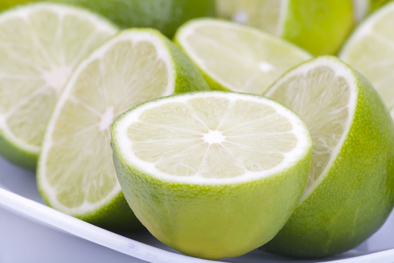 Close-up of several halved limes with their juicy interiors and green peels visible. The limes are placed on a white surface, with a few whole limes in the background, ready for use in a commercial citrus juicer. The image highlights the fresh and vibrant look of the citrus fruits.