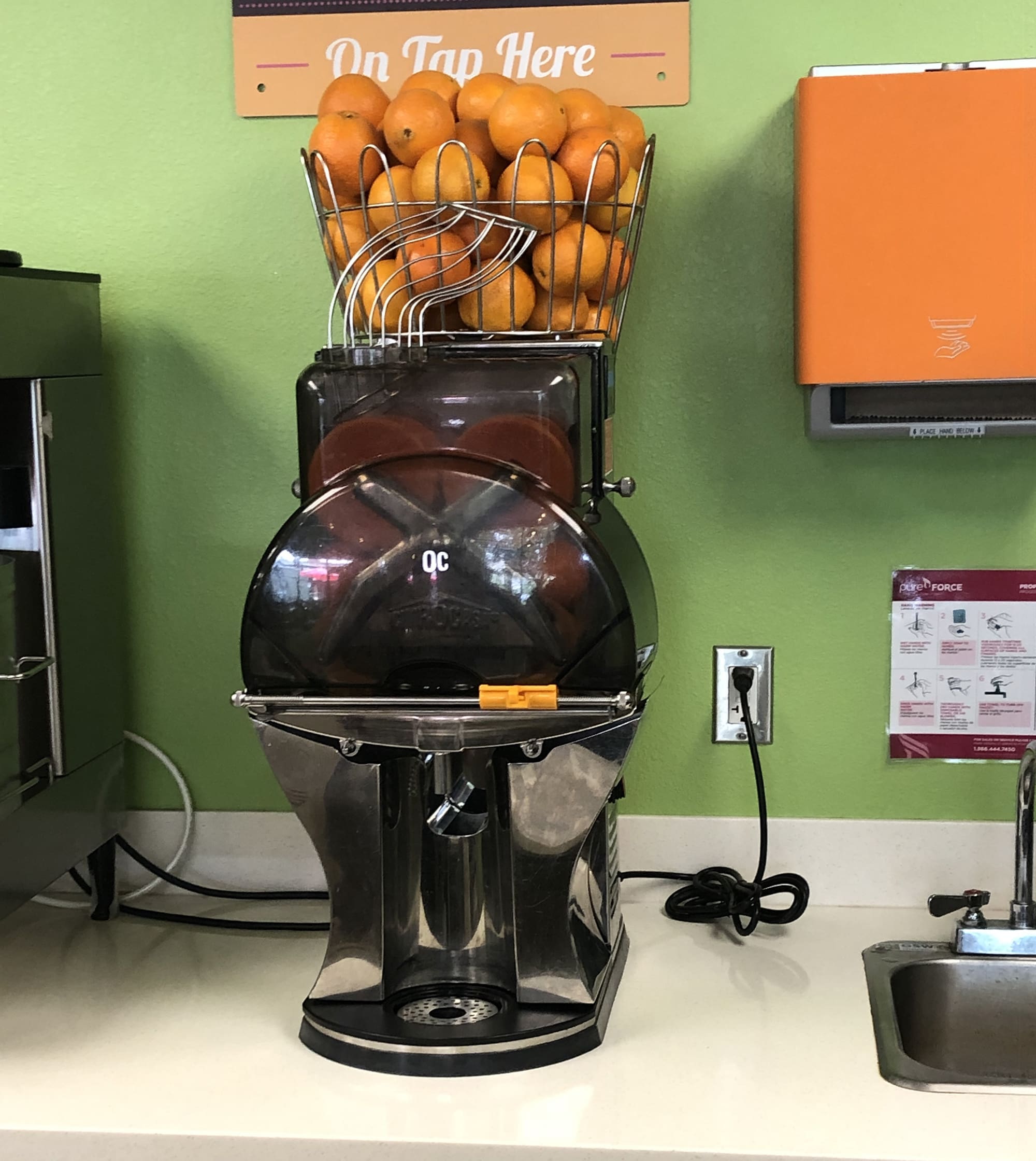 A commercial citrus juicer is placed on a countertop with a basket of fresh oranges on top. The appliance is surrounded by a green wall and an orange box mounted on the wall to its right. A small sink is visible to the right.