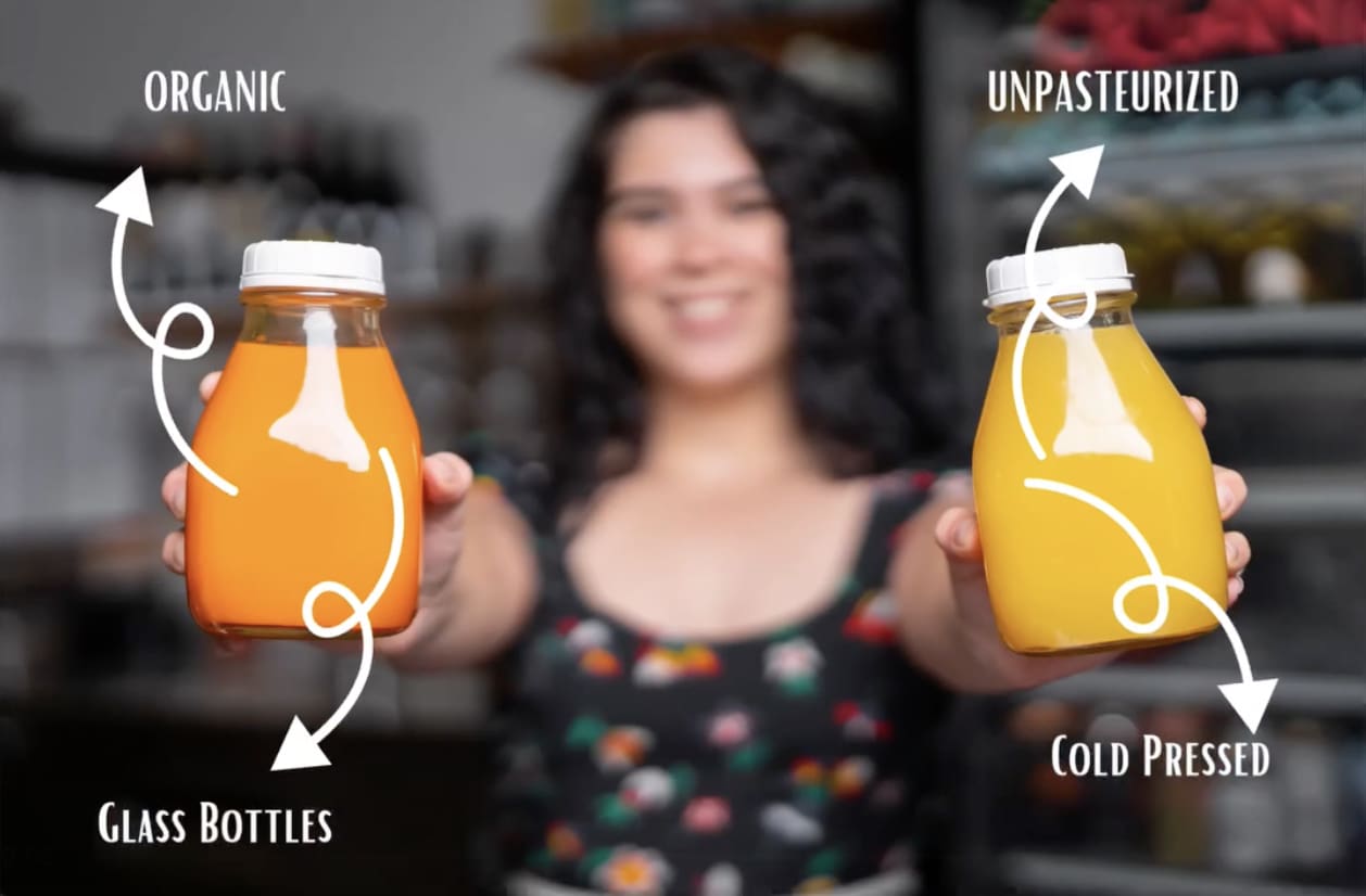 A smiling person holds two bottles of juice, one orange and the other yellow. Arrows and text point out that the juices, made with a commercial citrus juicer, are organic, in glass bottles, unpasteurized, and cold pressed. The person stands in front of shelves with various items.