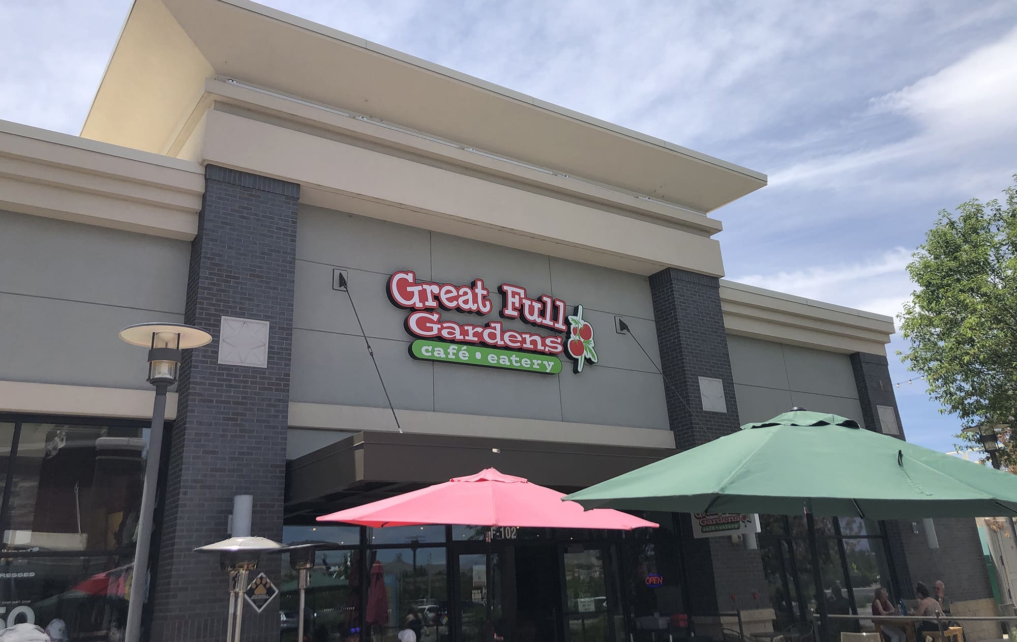 Exterior view of Great Full Gardens café and eatery. The building boasts a modern design with a sign above the entrance. Outdoor seating areas, with pink and green umbrellas, are visible in the foreground. A commercial citrus juicer sits enticingly near the entrance. The sky is partly cloudy.