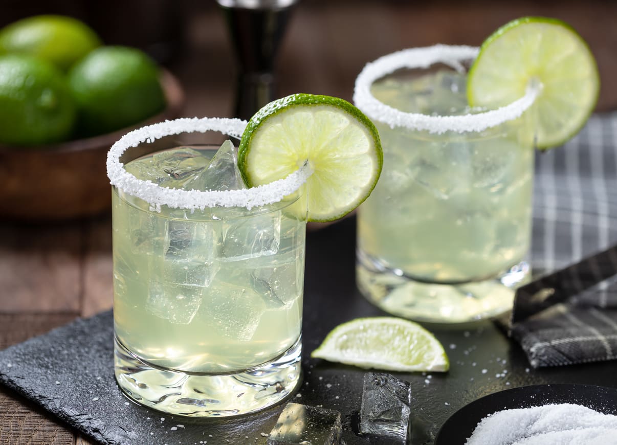 Two margarita glasses filled with ice and light green liquid sit on a black surface. The rims are coated with salt, and each glass is garnished with a lime wheel. A commercial citrus juicer stands in the background alongside a sliced lime and small bowl of salt.