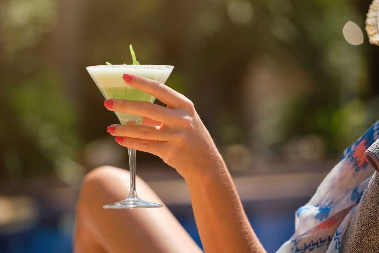 A person with red-painted nails holds a martini glass containing a green drink, garnished with a leaf, outdoors. The background is blurred, featuring lush greenery and warm lighting, evoking a relaxing, summery atmosphere—perfect for showcasing the fresh juices from our commercial citrus juicer.