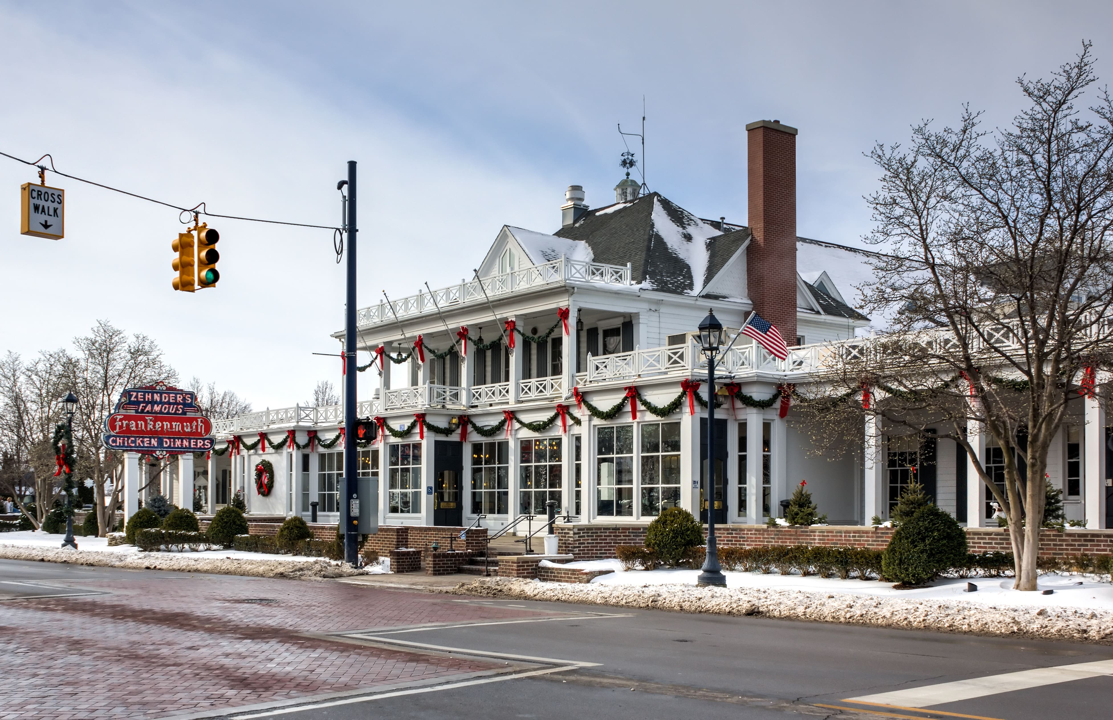 A two-story building decorated with Christmas garlands and wreaths features a sign that reads "Famous Chicken Dinners." The streets and sidewalks are lightly covered with snow, with bare trees, a commercial citrus juicer in the window, and traffic lights visible. An American flag is mounted on the balcony.