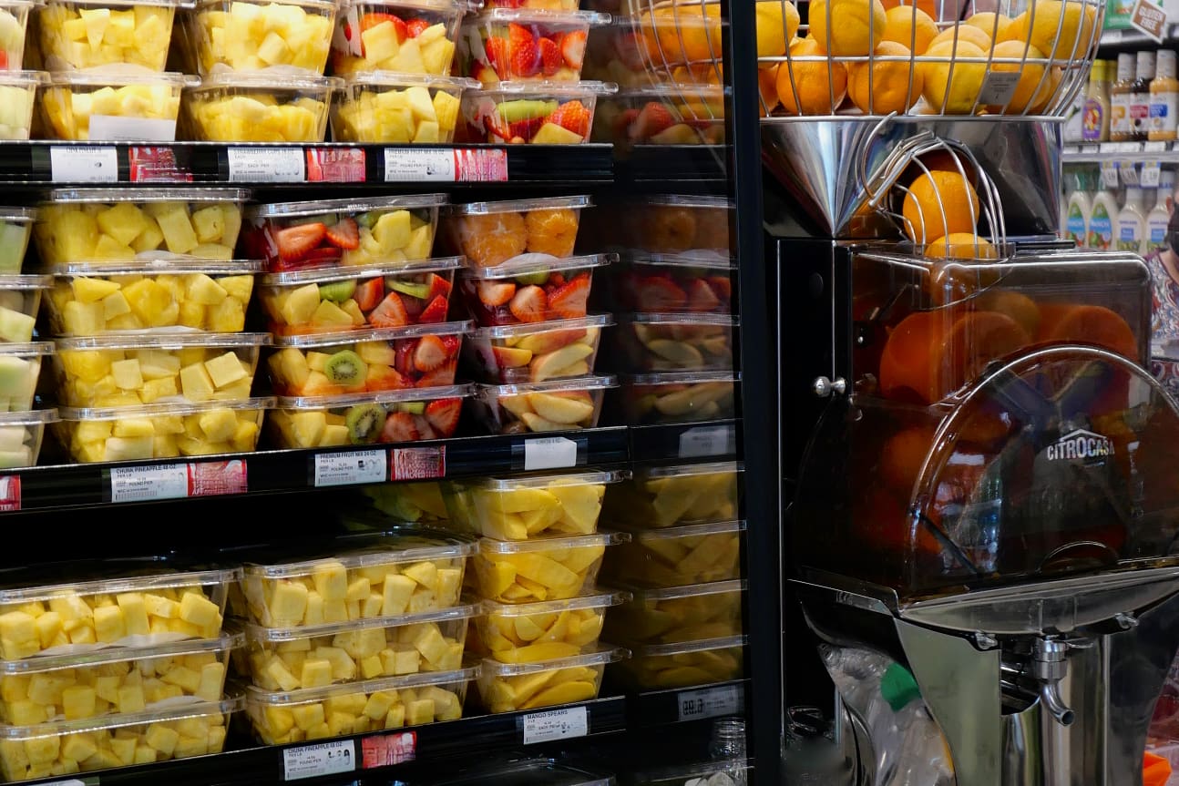 Shelves filled with plastic containers of pre-cut fruit, including pineapple, melon, kiwi, and strawberries, are displayed next to a commercial juicer. A basket of whole oranges sits on top of the machine in a grocery store setting.