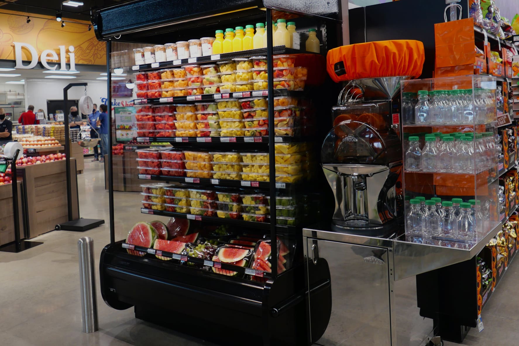 A grocery store aisle showcasing a refrigerated display filled with various pre-cut fruits in clear containers, including watermelon, cantaloupe, pineapple, and berries. Adjacent is a fresh juice dispenser by the commercial citrus juicer and shelves with bottles of water. The deli section is visible in the background.