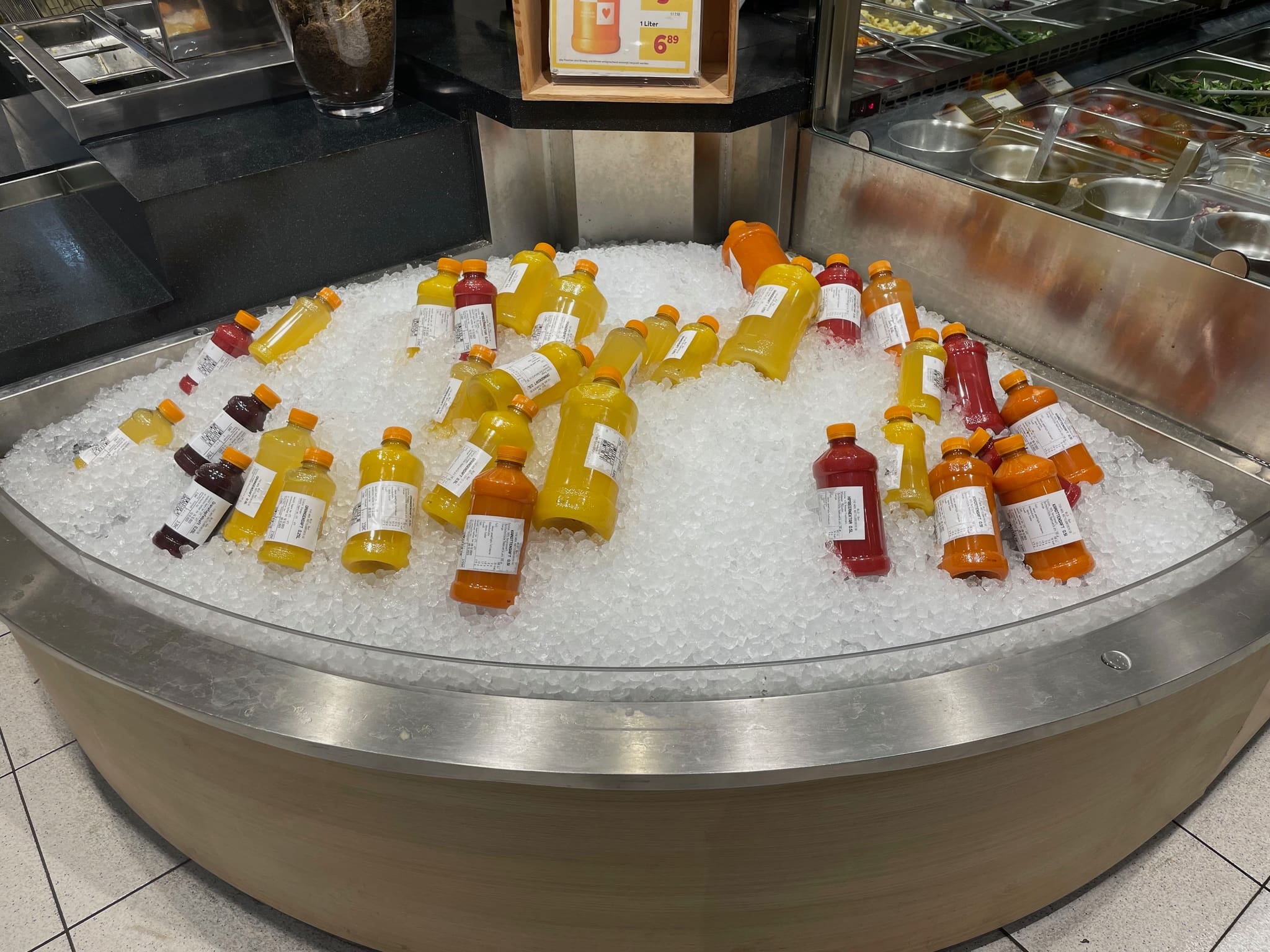 A refrigerated display filled with various bottles of juice, freshly squeezed by a commercial citrus juicer, rests on a bed of ice. The bottles come in different flavors, glowing in vibrant shades of orange, yellow, pink, and red. In the background, part of a salad bar with trays of ingredients can be seen.