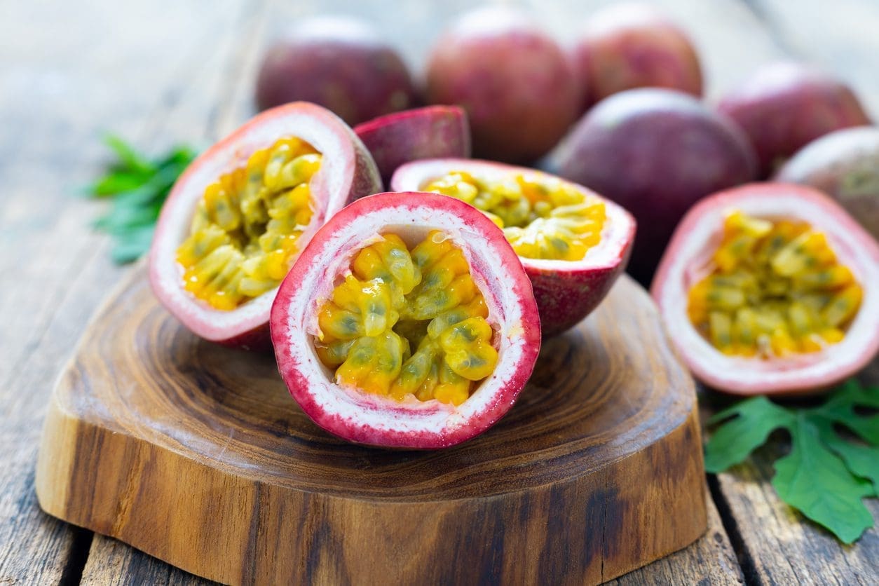 Sliced passion fruits with vibrant yellow and orange pulp displayed on a wooden board, perfect for crafting the best juice recipe. Whole passion fruits are in the background, slightly out of focus. The board rests on a rustic wooden surface, and a few green leaves are scattered around.