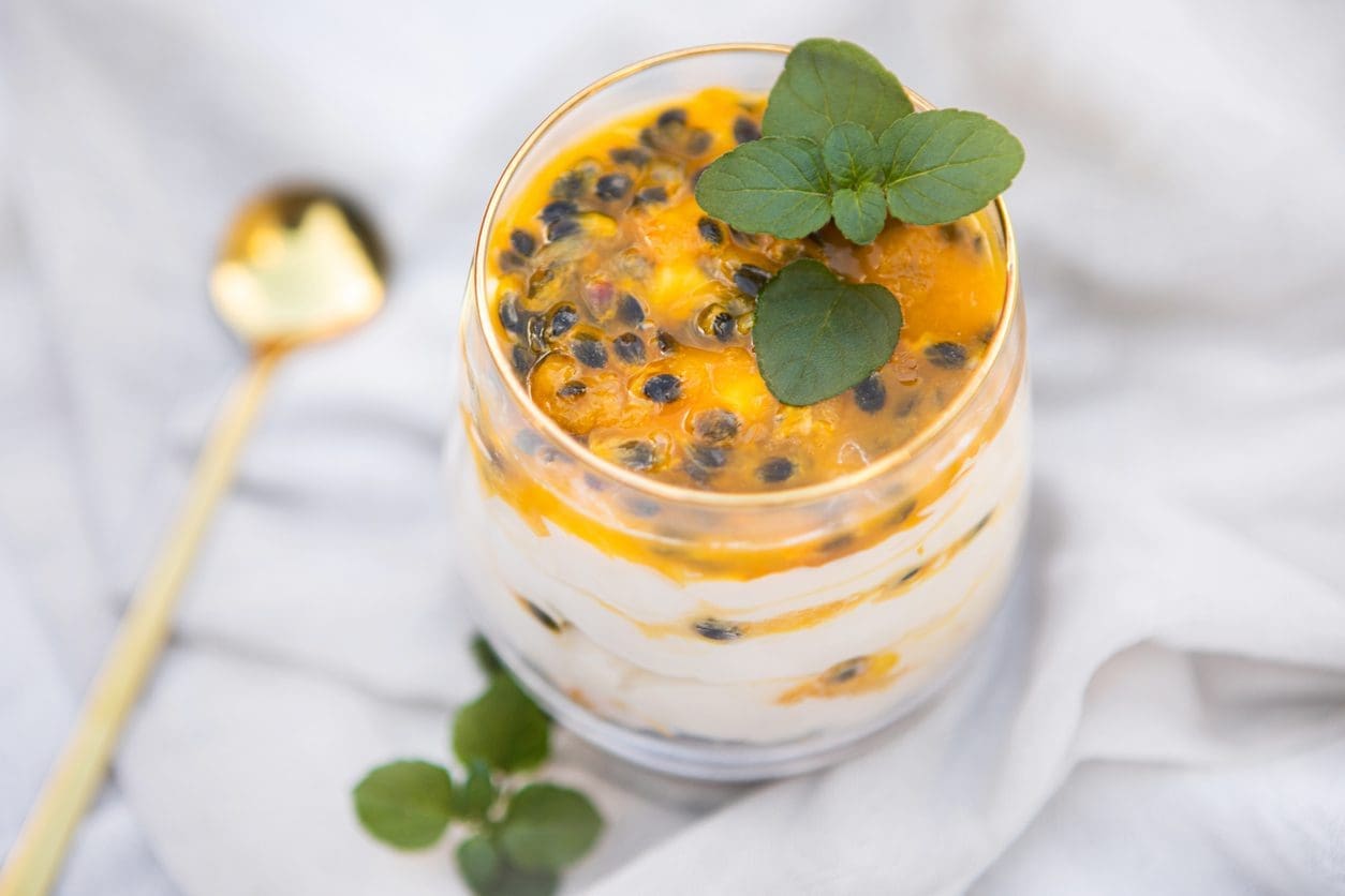 A glass is filled with layers of creamy yogurt and homemade passion fruit, topped with fresh passion fruit pulp and garnished with a sprig of mint. A golden spoon lies on a light fabric next to it, making this an exquisite part of any Passion Fruit Juice Recipe collection.