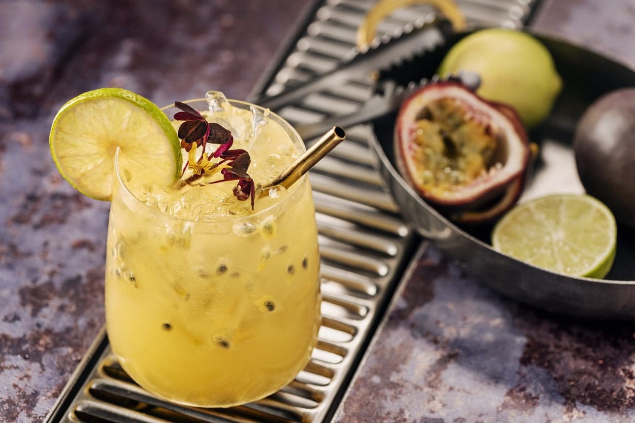 A refreshing cocktail garnished with a lime slice and fresh herbs in a glass with a metal straw sits on a metal surface. Beside it, there's a halved passion fruit, a whole passion fruit, and a halved lime in a bowl. Discover the best passion fruit juice recipe for enhancing your homemade drinks.