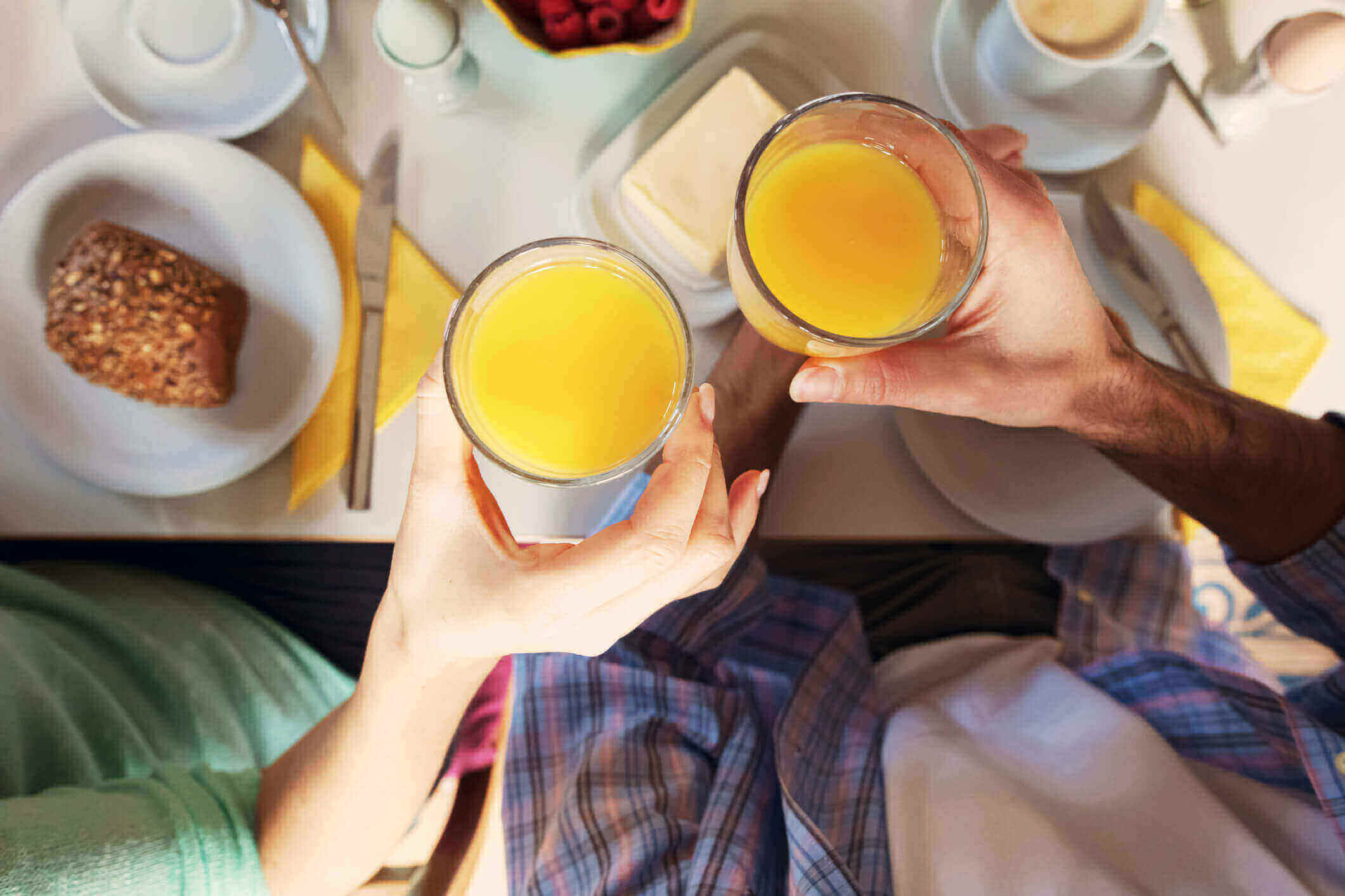 Two people sit across from each other at a breakfast table at Great Full Gardens. They clink glasses of orange juice above a spread of bread, berries, butter, and coffee. The table is laid with light blue plates and utensils.