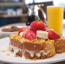 A close-up of a breakfast dish at Great Full Gardens featuring two thick slices of French toast topped with fresh strawberries and banana slices. The toast is drizzled with creamy white sauce and sprinkled with crushed cereal. A glass of orange juice and a coffee pot are in the background.