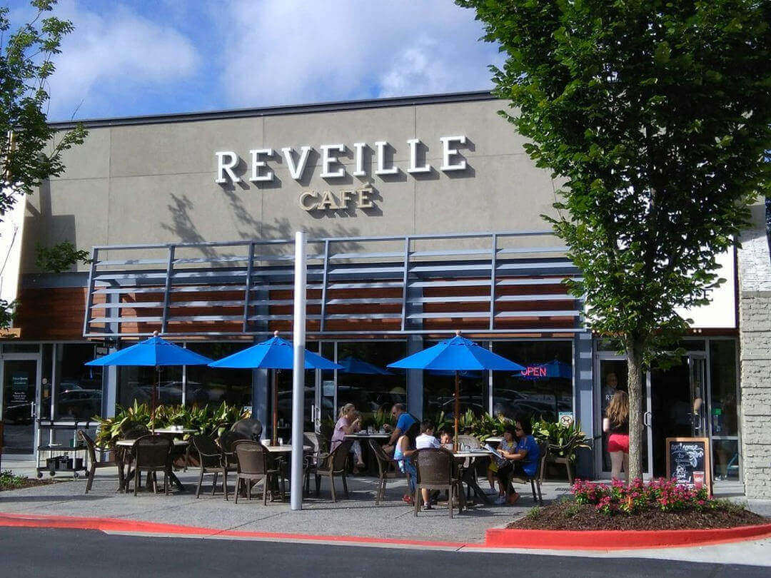 Outdoor seating area of Reveille Café, featuring several tables with people dining under blue umbrellas. The café has a modern facade with large windows, and there's greenery decorating the space. A sign on the building displays the café's name, reminiscent of the charming ambiance at Great Full Gardens.