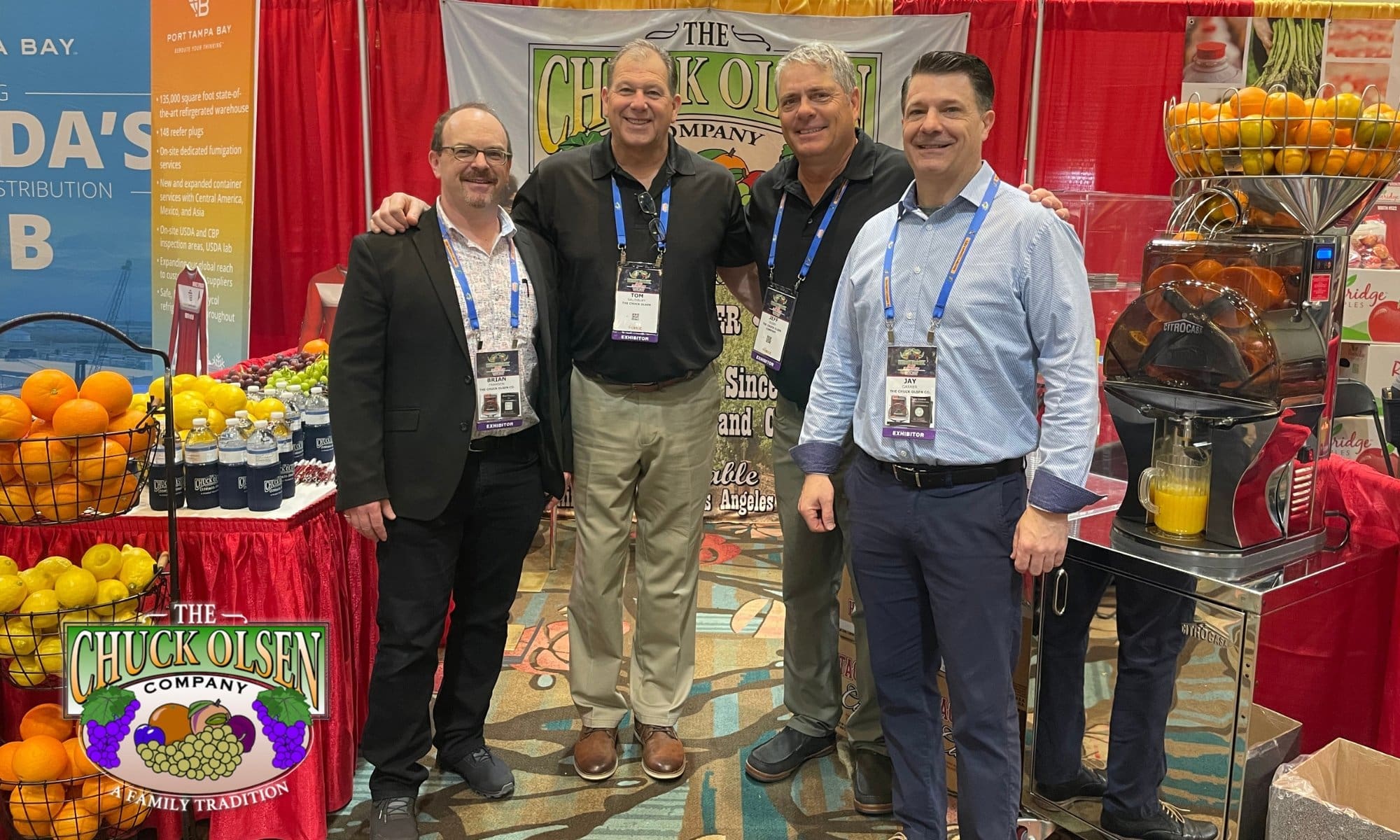 Four men stand smiling in front of a booth with a Chuck Olsen Company banner at the SEPC Show 2025. They are surrounded by baskets of oranges and displays of orange juice. The background features red curtains and promotional materials.