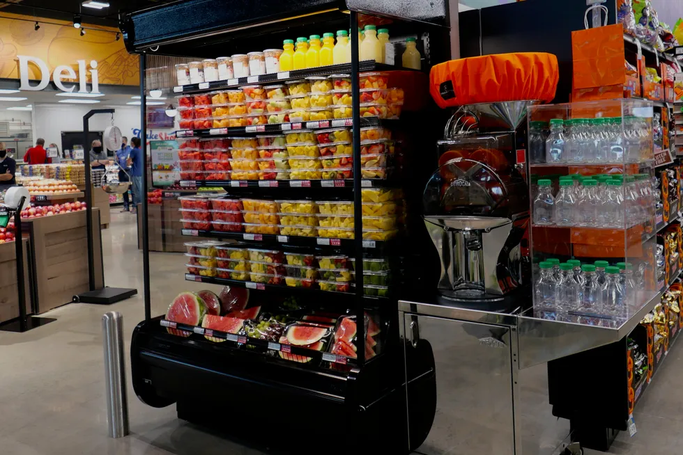 A grocery store display with shelves of assorted pre-cut fruit containers, bottled citrus juice, a juicing machine, and bottled water. In the background, there is a deli section and produce area with apples.