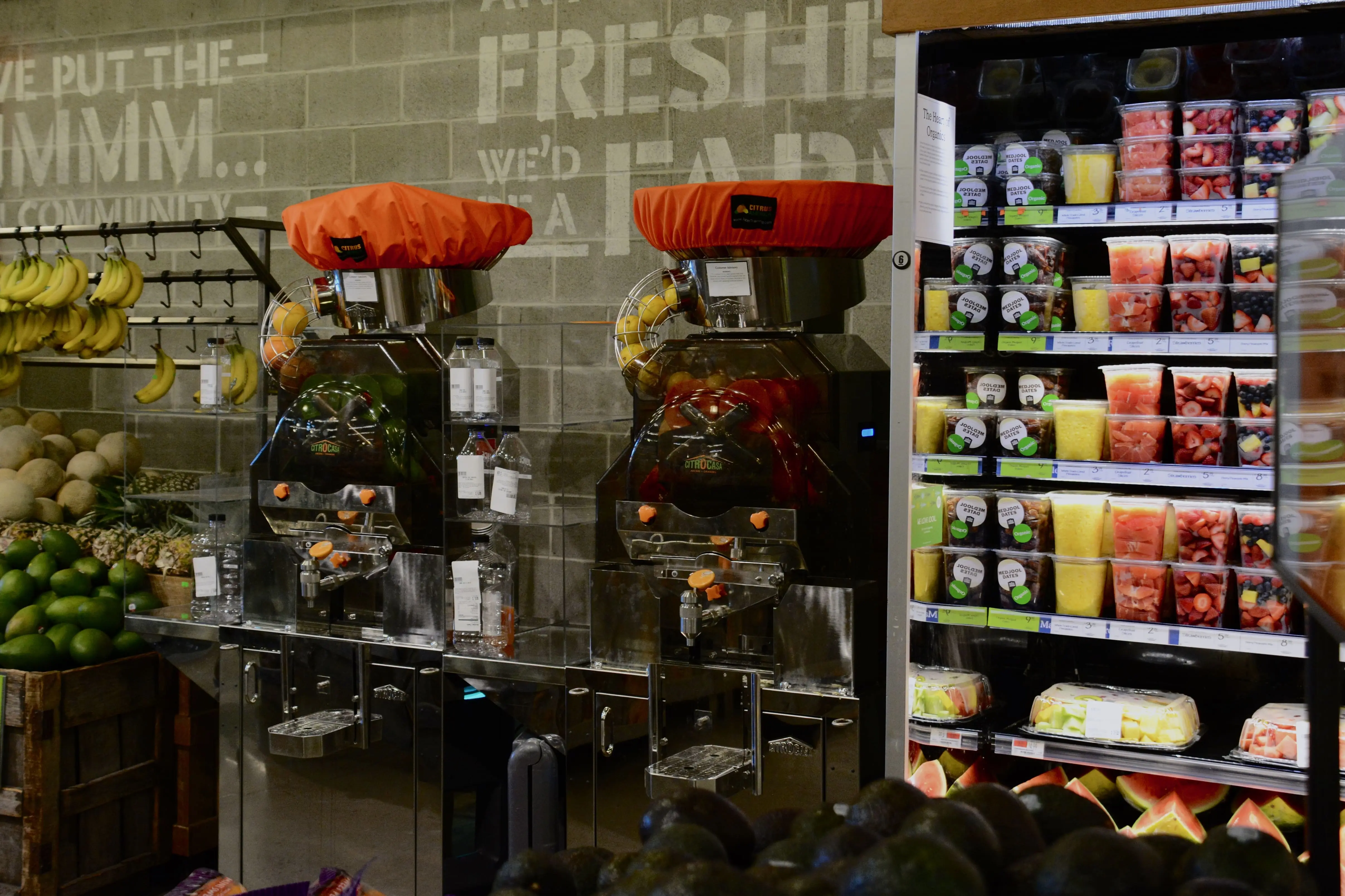 Self-serve orange juice machines next to a refrigerated display of pre-cut fruit containers in a grocery store. Bananas and avocados are visible on the left side.