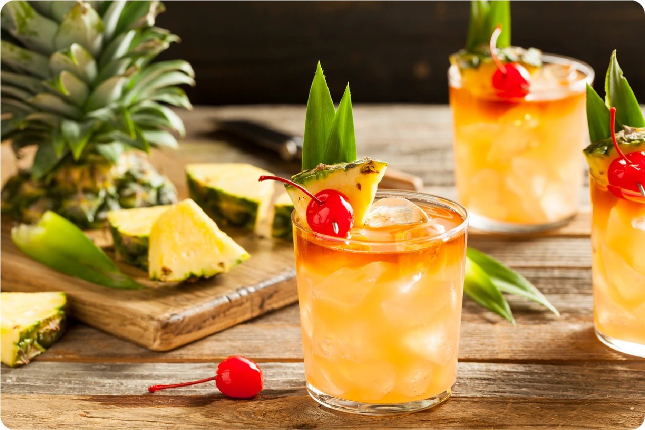 A glass of tropical cocktail garnished with a pineapple slice, green leaves, and a cherry, sits on a wooden table with more drinks and fresh pineapple pieces in the background.
