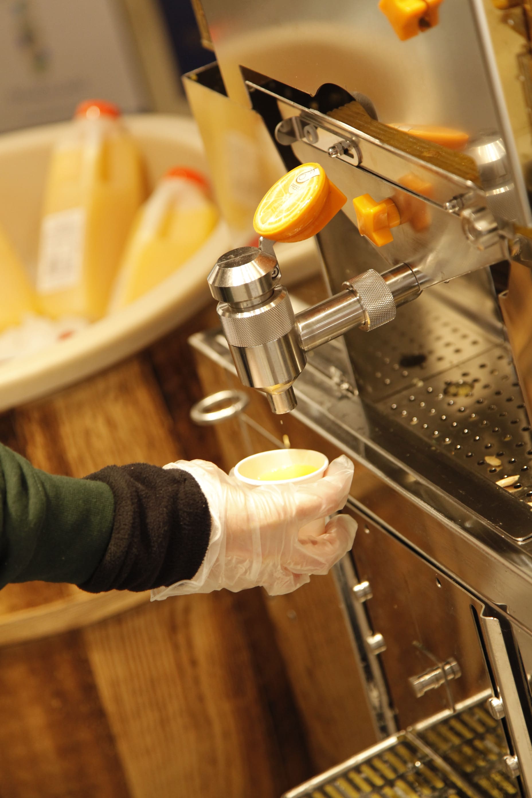 A gloved hand holds a small cup under a metal dispenser, filling it with orange juice at a self-serve machine in Barons Market. Yellow bottles are visible in the background.