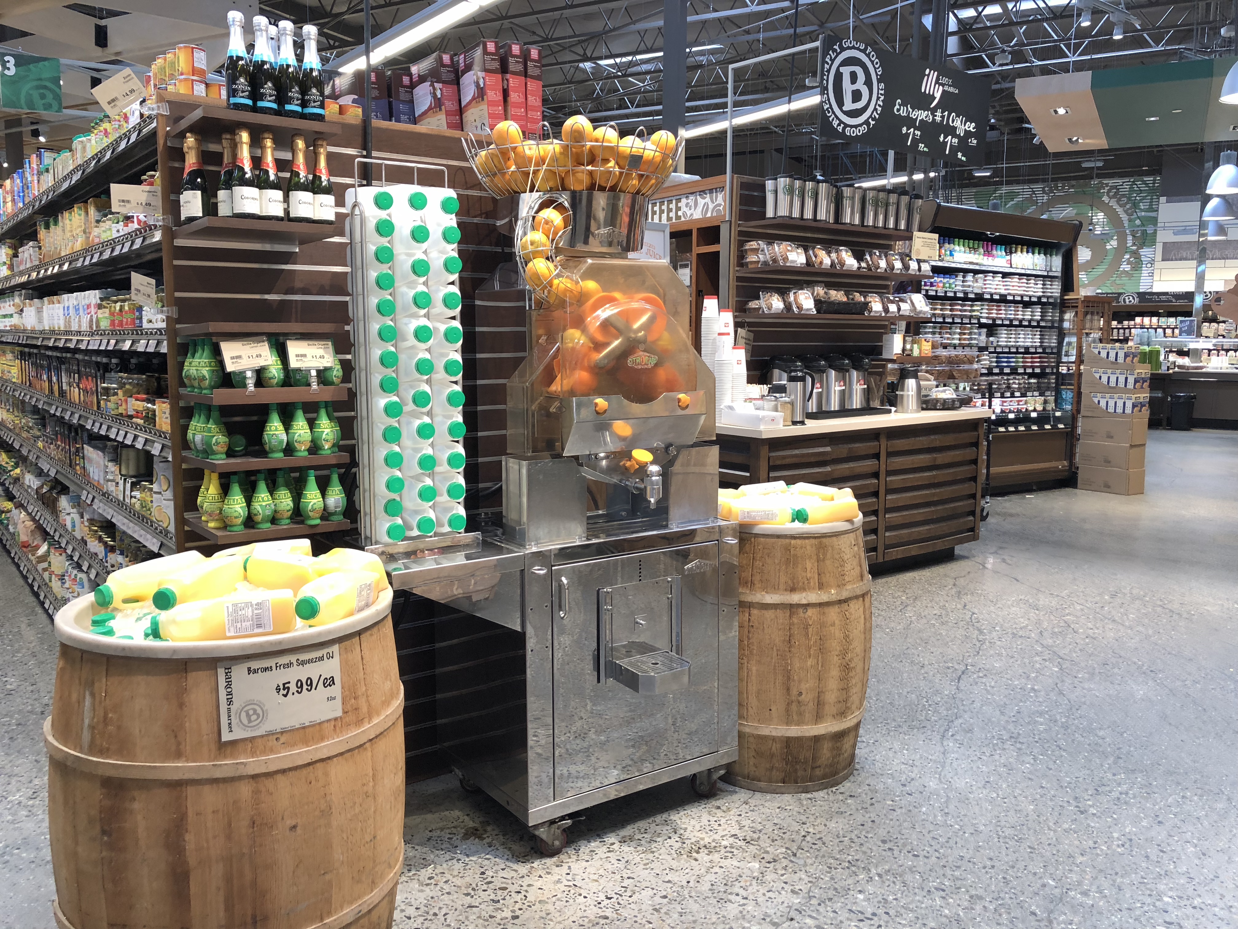 At Barons Market, a Citrus America orange juice machine sits topped with fresh oranges. Bottles and cartons of juice rest on wooden barrels, while shelves stocked with groceries provide a vibrant backdrop.