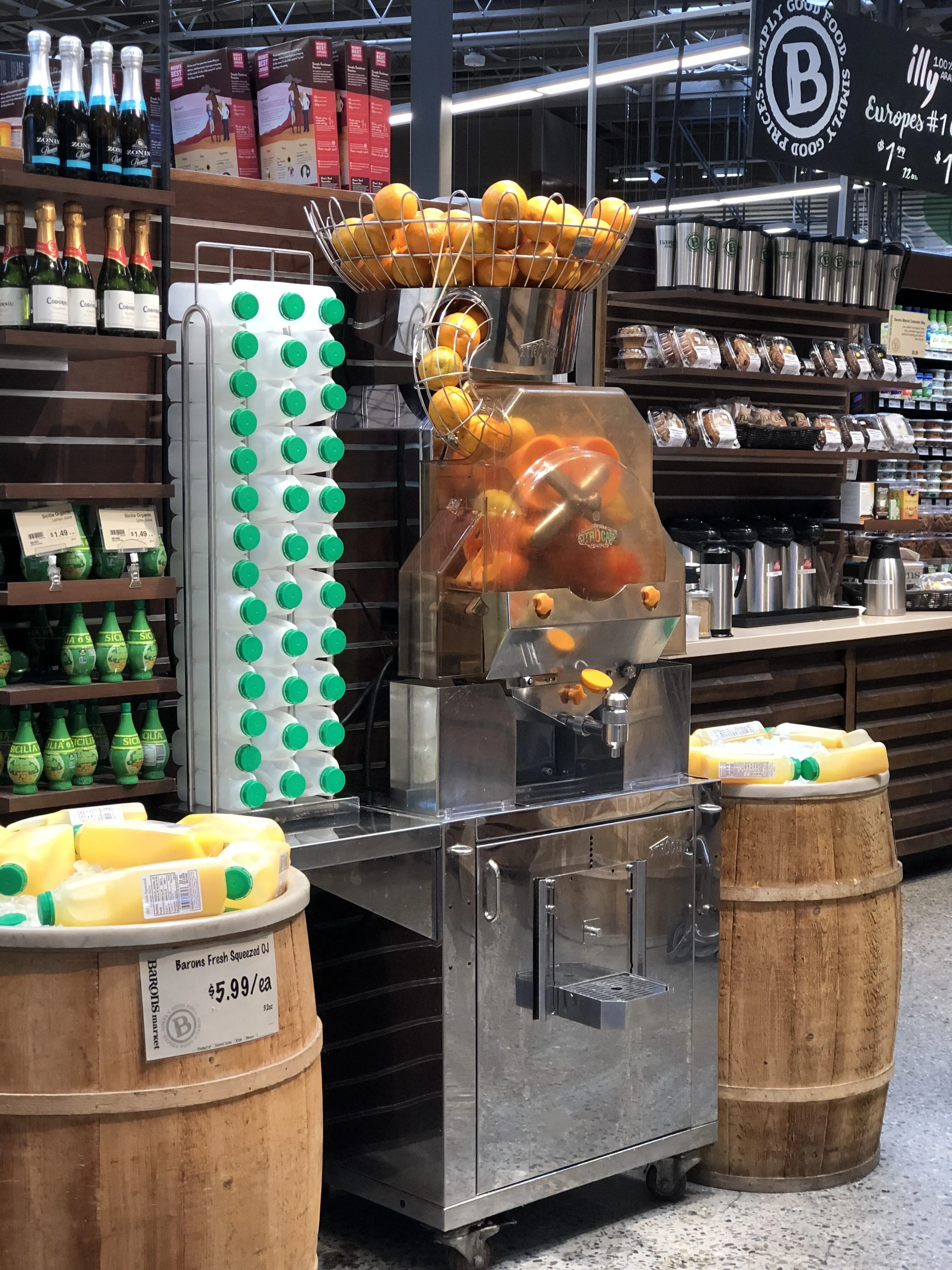 A grocery store orange juicing machine with oranges stacked on top, fresh juice flowing into bottles, and shelves of juice bottles and other products nearby.