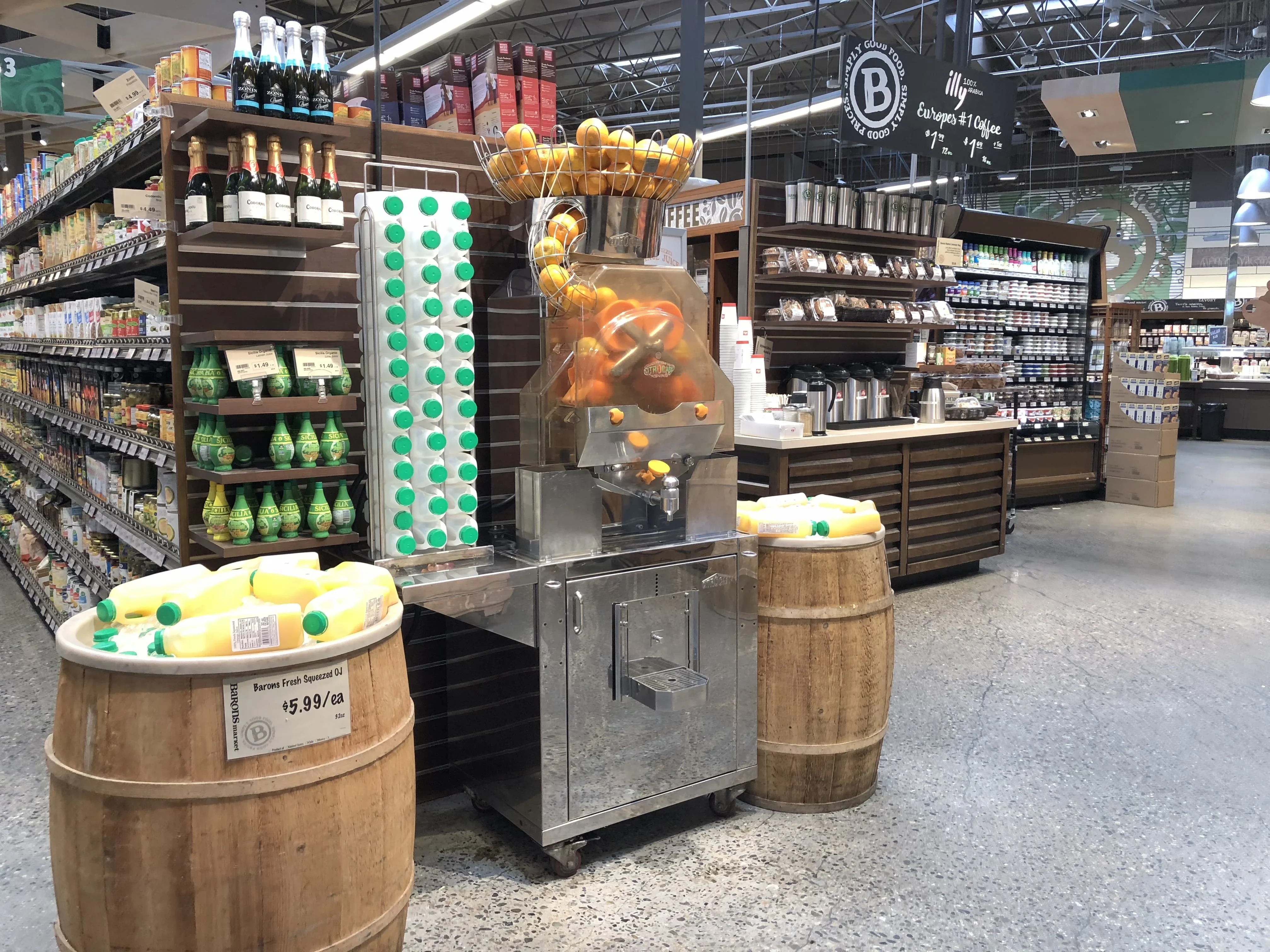 A grocery store features a self-serve orange juice machine with whole oranges inside, surrounded by bottled juice on wooden barrels and shelves stocked with groceries and beverages in the background.