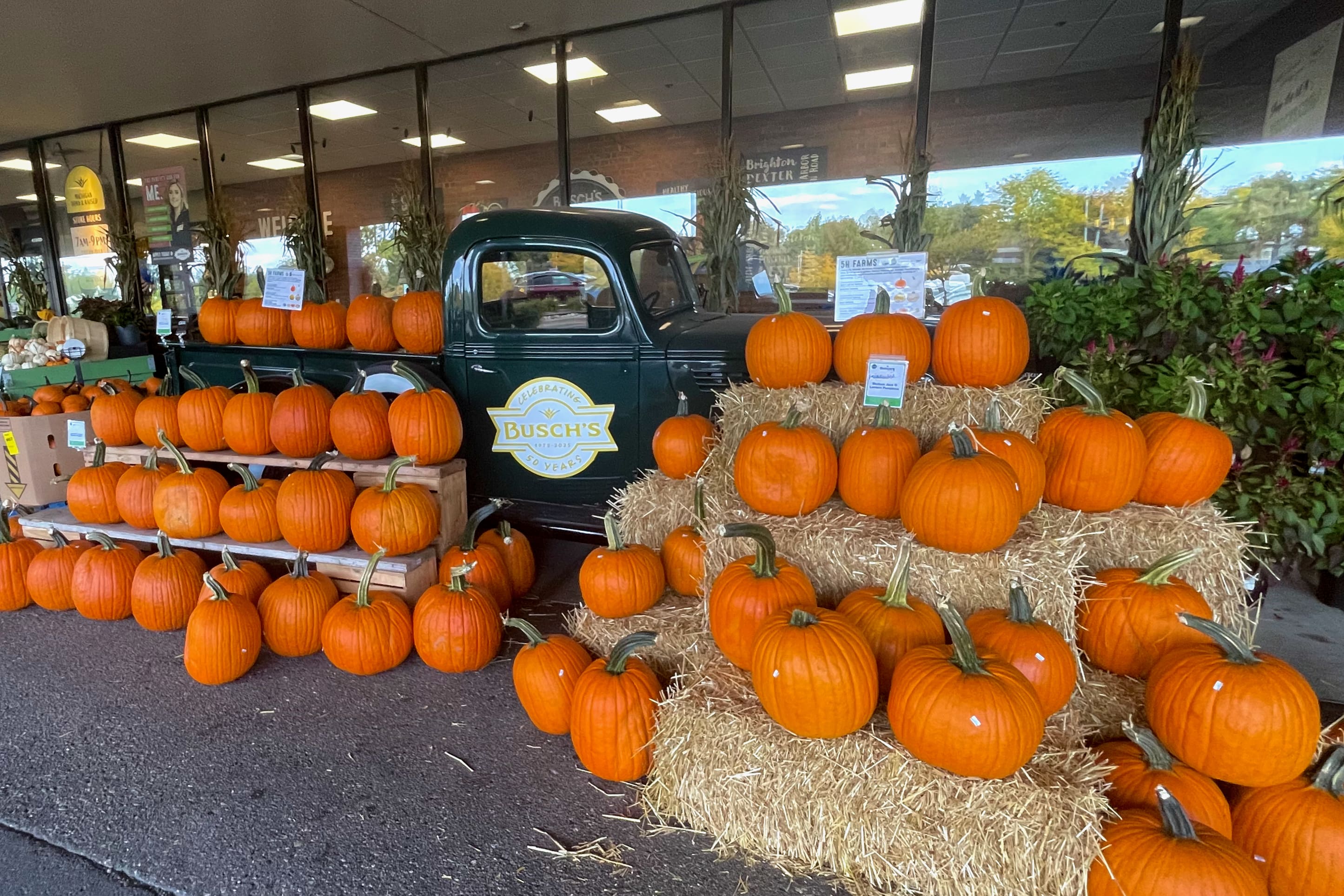 A vintage green truck is parked outside a store, surrounded by neatly stacked bright orange pumpkins and bottles of Barons Market orange juice on hay bales and wooden shelves, creating a festive autumn display.