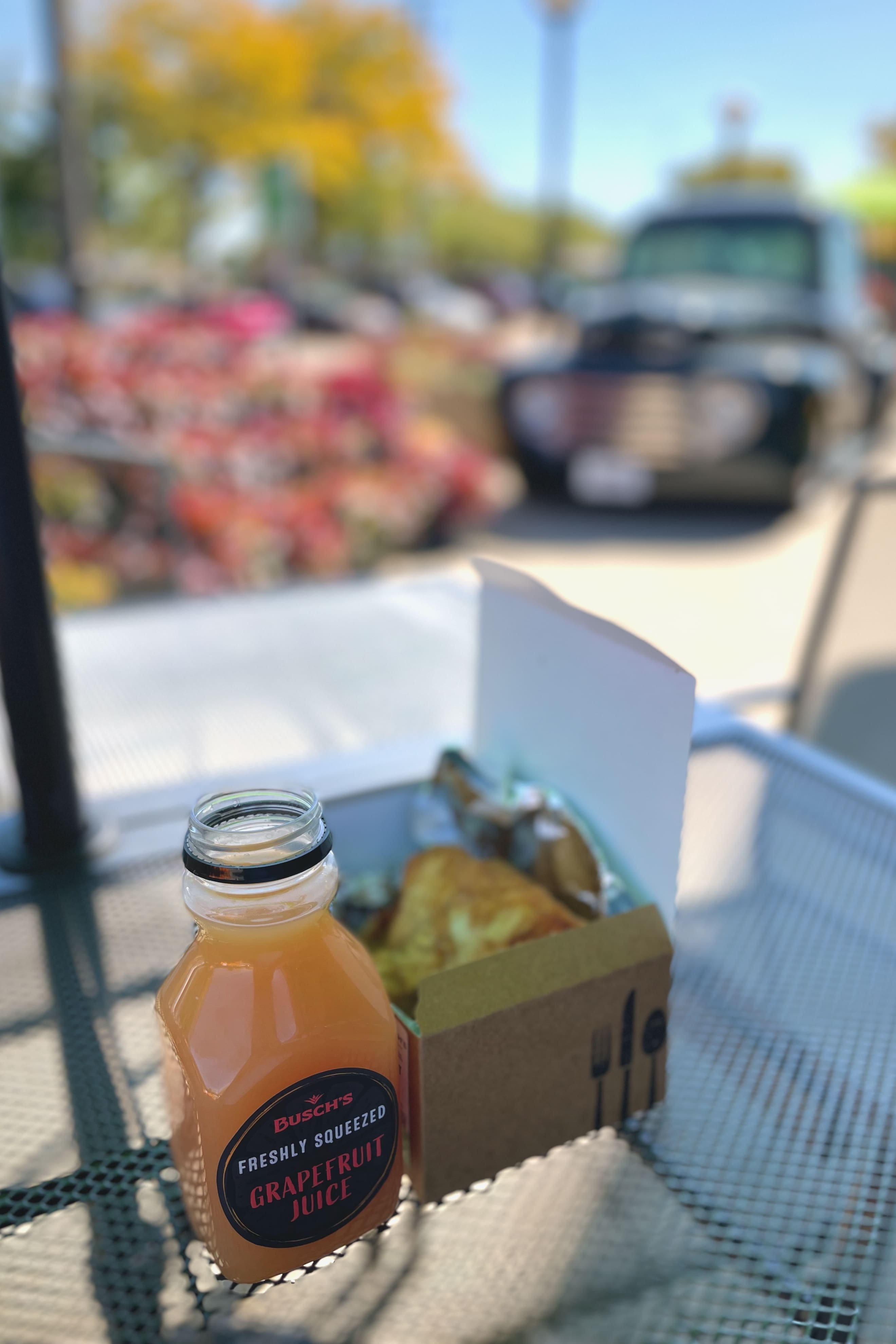 A bottle from the fresh juicing program and a boxed meal sit on a metal outdoor table, with colorful flowers and a black food truck visible in the blurred background.