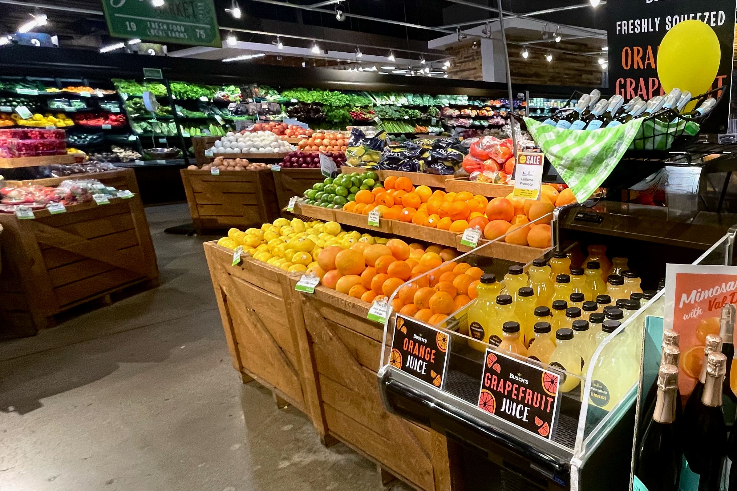A grocery store produce section displays colorful fruits, including oranges, lemons, and apples, with bottles of orange and grapefruit juice from the fresh juicing program in the foreground and various vegetables in the background.