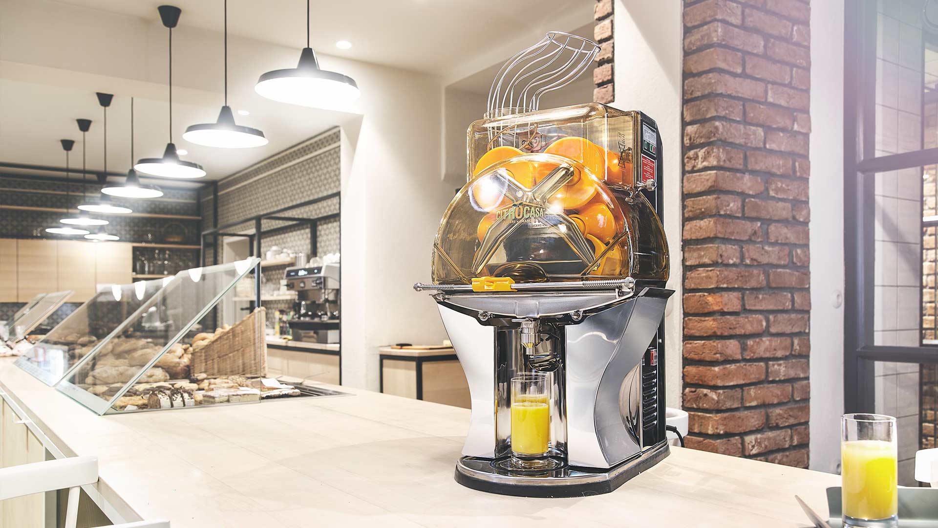 A commercial citrus juicer squeezes fresh oranges, dispensing juice into a glass on a countertop in a modern, well-lit café with brick and glass details.