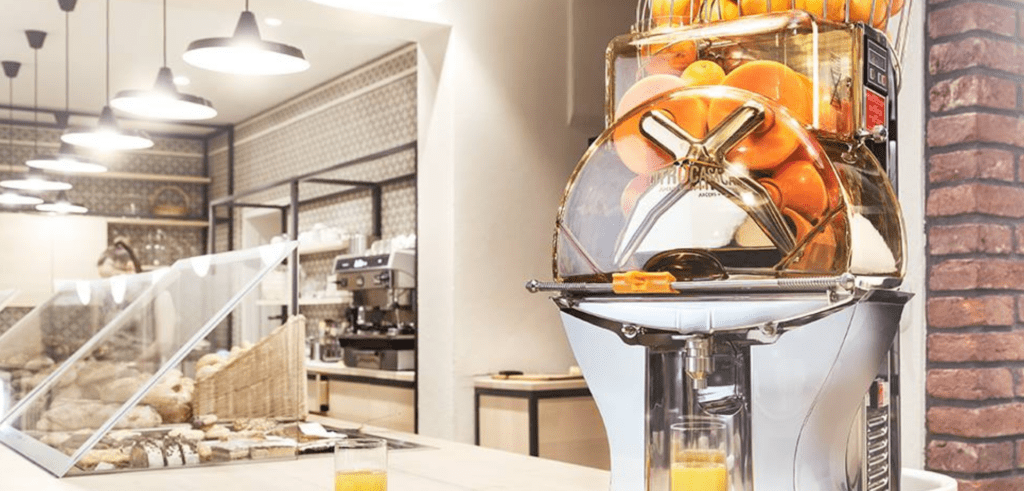 A modern café counter features a citrus juicer filled with oranges, dispensing fresh orange juice into a glass. Nearby are pastries in baskets, a coffee machine, and hanging lights illuminating the space.
