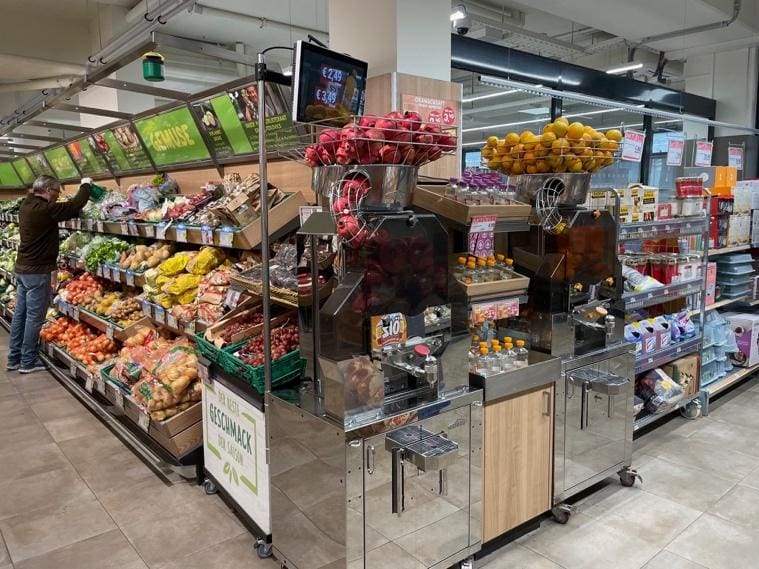 A supermarket produce section displays fruits and vegetables on shelves; in the foreground, a Commercial Citrus Juicer with oranges and pomegranates on top dispenses fresh juice into bottles. A person is selecting produce on the left.