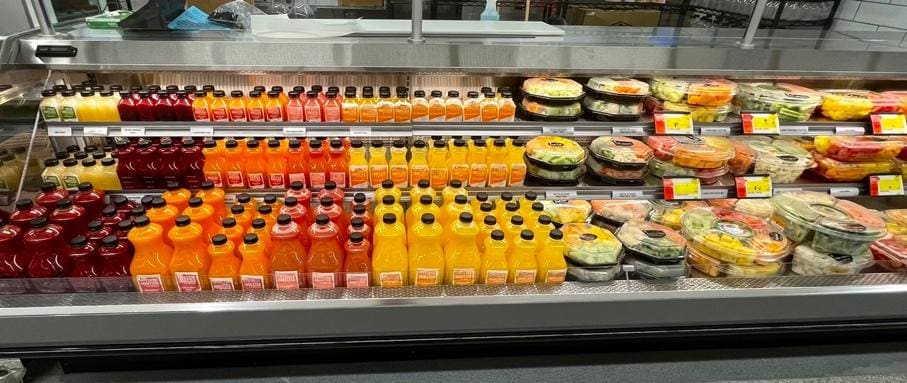 Refrigerated grocery display with rows of assorted juice bottles in red, orange, and yellow—some featuring juice made fresh with a commercial citrus juicer—alongside various packaged salads, fruit bowls, and deli items on upper shelves.