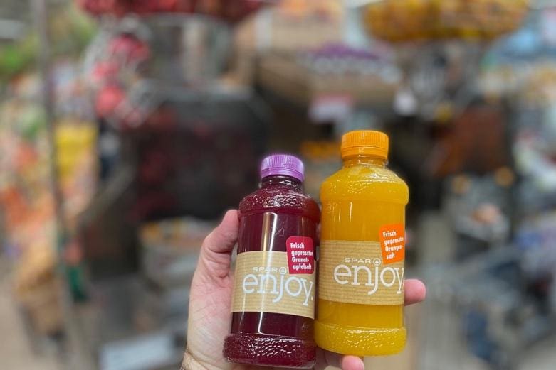A hand holds two SPAR enjoy juice bottles in a grocery store—one with a red drink, the other orange, both likely crafted using a Commercial Citrus Juicer. Blurred shelves create a vibrant backdrop.