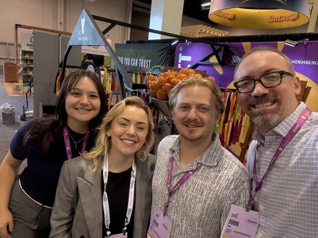 Four people smiling and posing for a selfie at an indoor event or convention. They are wearing name badges and standing in front of a colorful booth with banners and a fruit display, celebrating the fresh revolution in produce.