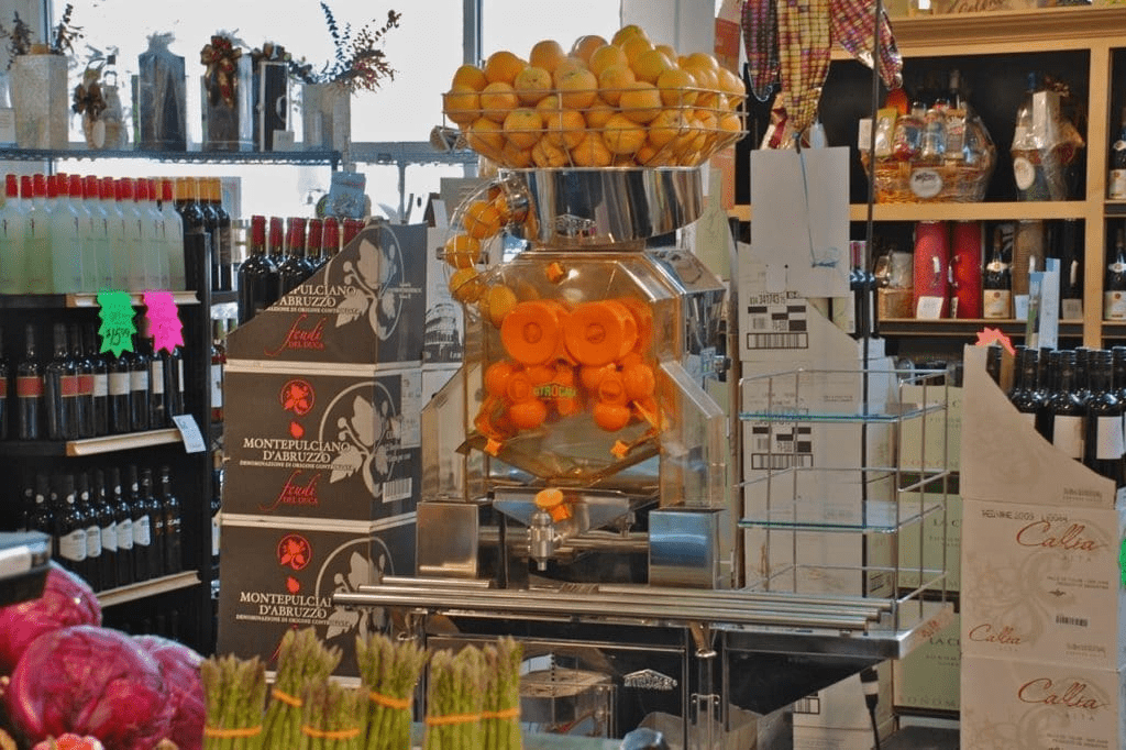 A grocery store display features a Commercial Citrus Juicer filled with oranges and a bowl of lemons on top. Wine bottles, boxes, produce, and various food items are arranged around the juicer.