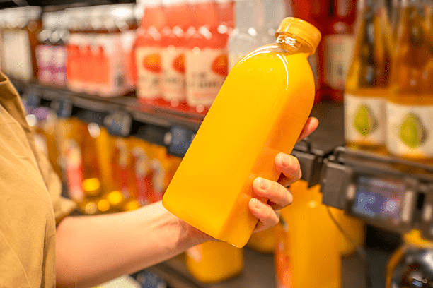 A person holds a bright yellow bottle of juice in a grocery store aisle, surrounded by colorful drink bottles—freshly squeezed, perhaps with a commercial citrus juicer, for vibrant flavor and appeal.